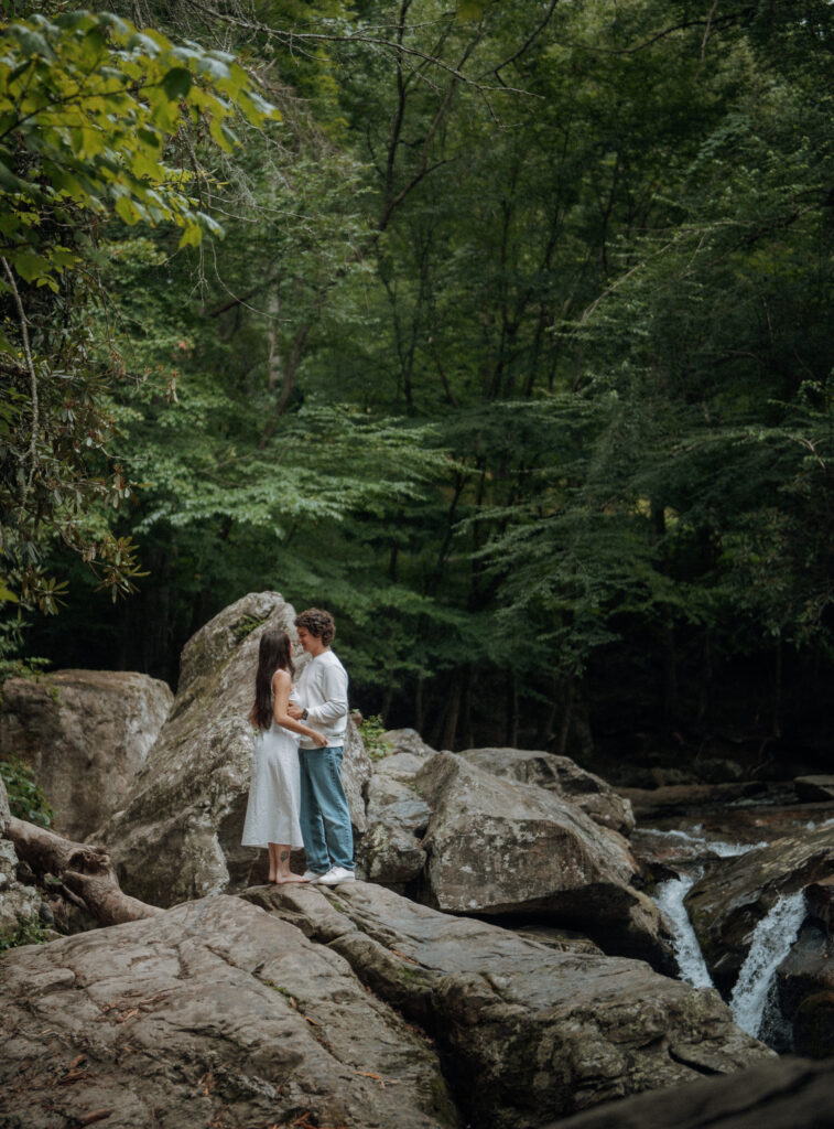 Destination engagement photos at Trash Can Falls in the Blue Ridge Mountains, with overcast skies, lush forest, and romantic waterfall backdrops. Captured by destination wedding and engagement photographer KNCAPTURES. Couple standing in front of waterfall.