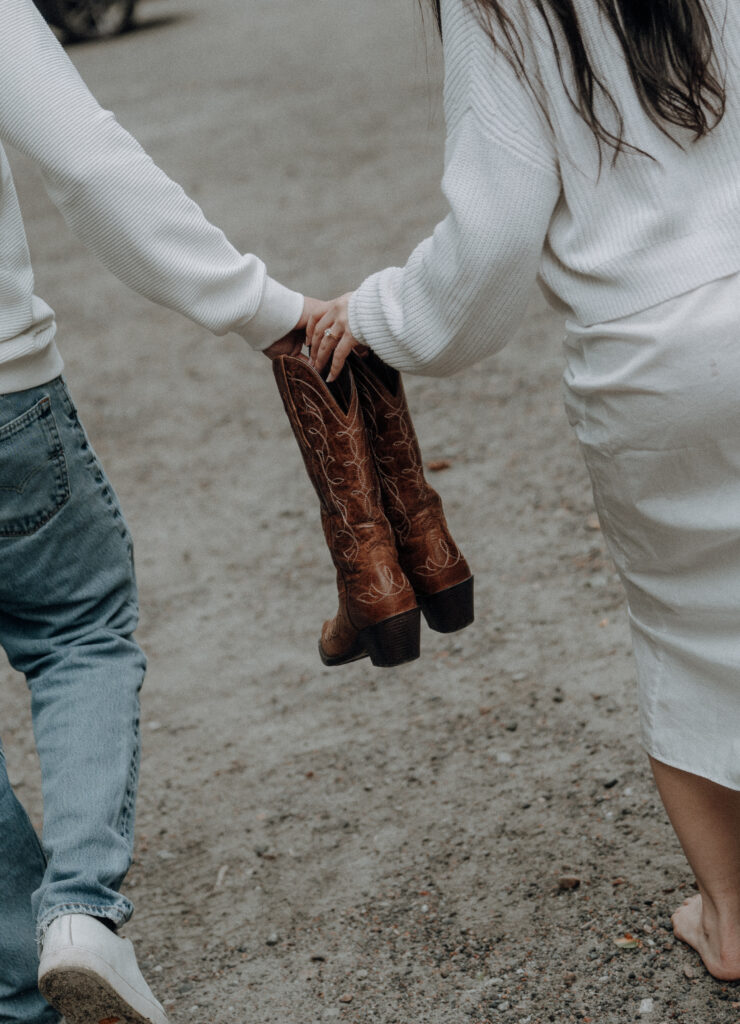Destination engagement photos at Trash Can Falls in the Blue Ridge Mountains, with overcast skies, lush forest, and romantic waterfall backdrops. Captured by destination wedding and engagement photographer KNCAPTURES. Couple holding a pair of cowboy boots