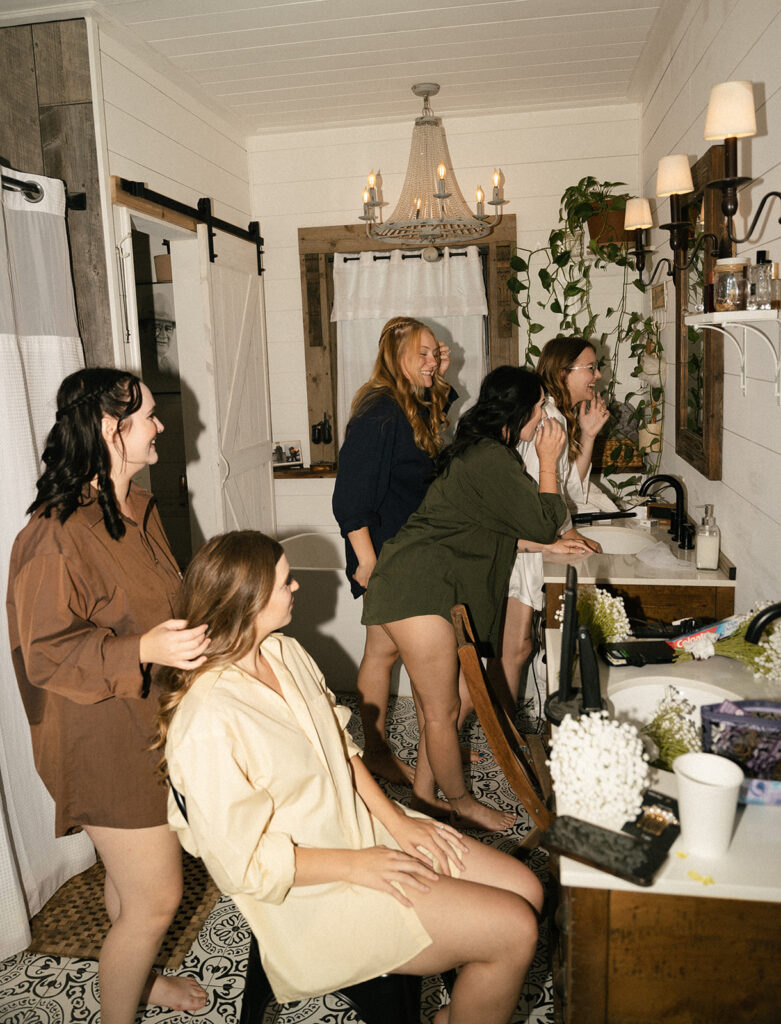 getting ready photos of bridesmaids in matching button up shirts doing their hair and makeup 